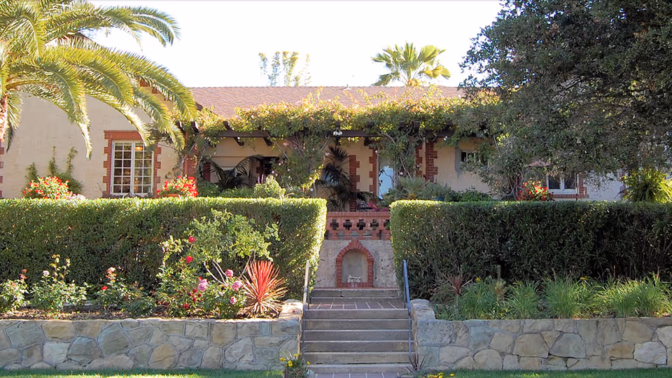 Front exterior view of a single-story building with a stone retaining wall, steps leading up to a porch covered with greenery, surrounded by trimmed hedges, flowering plants, and palm trees.