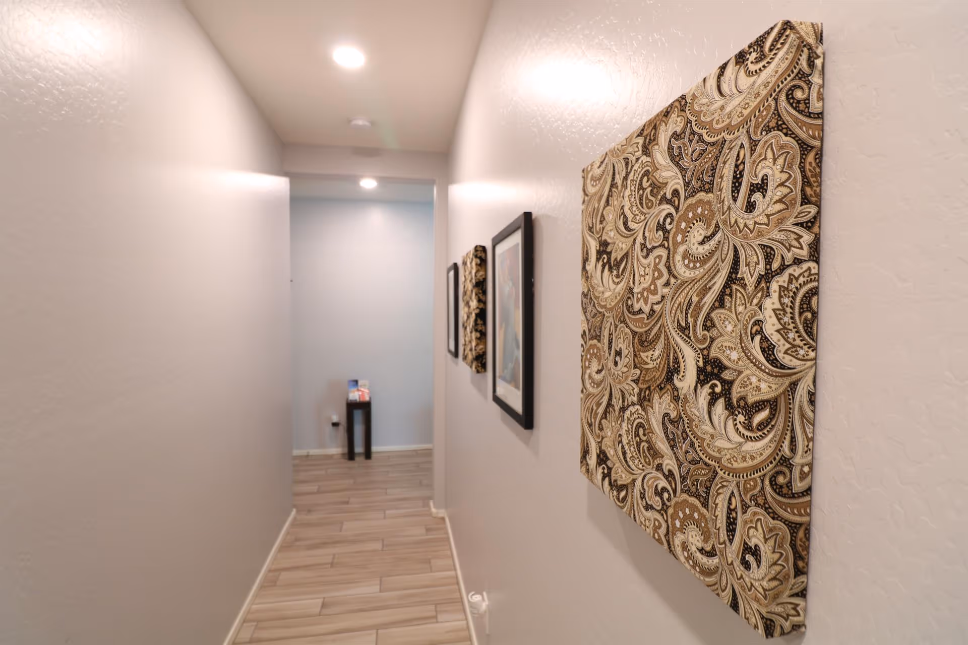 A narrow hallway with light-colored walls and wood-patterned tile flooring. The hallway is decorated with framed artwork and a patterned fabric wall hanging on the right side. At the end of the hallway, there is a small black table with some items on it. The ceiling has recessed lighting.