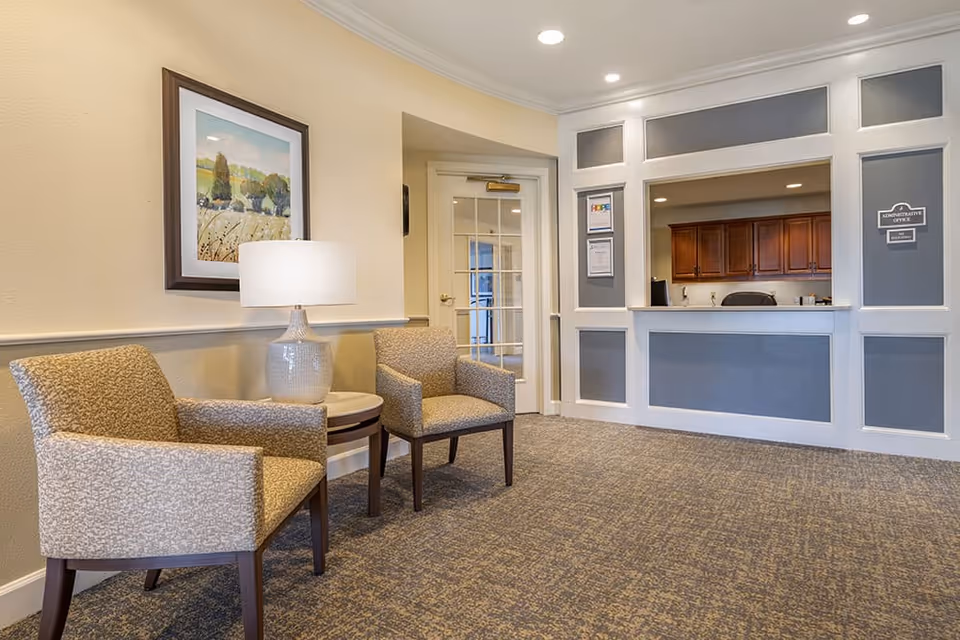 A reception area with two patterned armchairs and a small round table with a lamp between them. Behind the reception desk, there are wooden cabinets and a sign that reads 'Administrative Office'. The walls are painted beige with white and gray paneling, and there is a framed landscape painting on the wall.