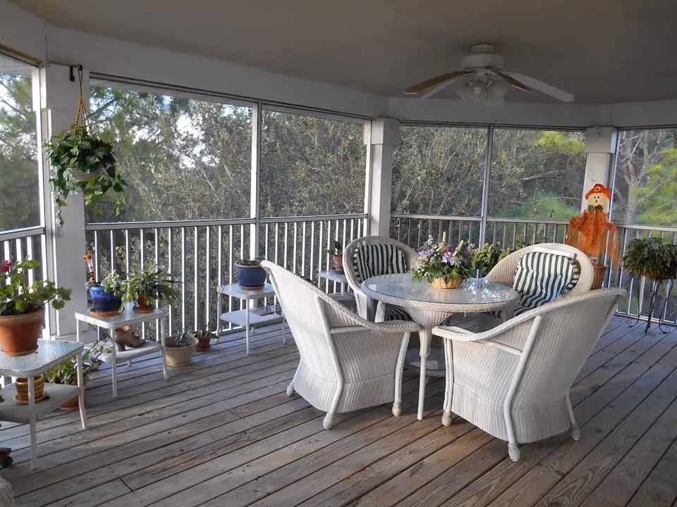 Screened-in wooden porch with a round glass-top table, four white wicker chairs with striped cushions, and several potted plants.