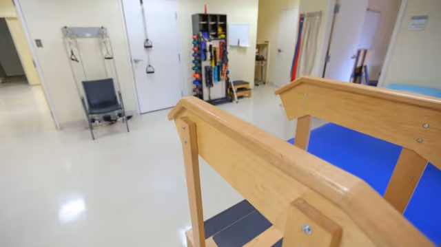 Interior view of a rehabilitation or physical therapy room with wooden parallel bars for walking exercises, a chair with straps, and a rack holding various exercise equipment such as dumbbells and resistance bands.