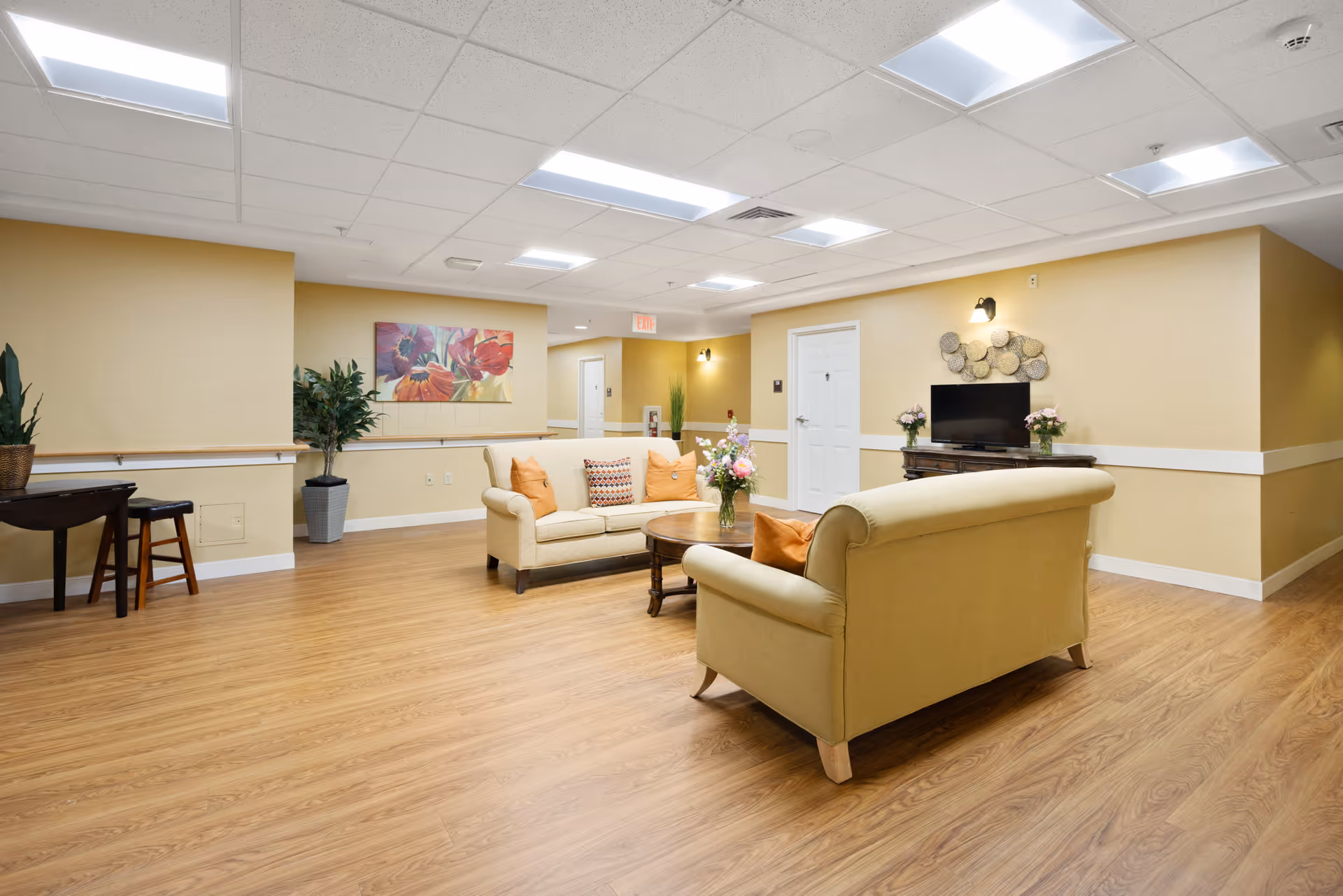 A bright common lounge with two beige sofas around a coffee table, a TV on a console, plants, artwork, and wood-look flooring.