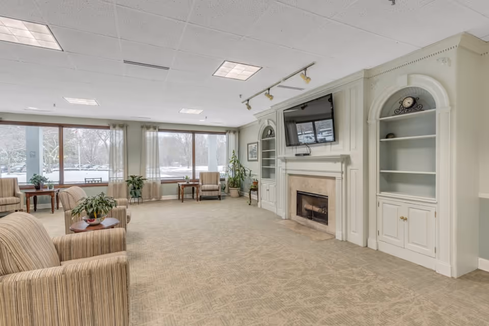 A spacious living room area with large windows letting in natural light. The room features beige carpet, several striped upholstered armchairs, small wooden tables with plants, and built-in white cabinetry with a fireplace and a mounted flat-screen TV above it. There are also some decorative items on the shelves and track lighting on the ceiling.