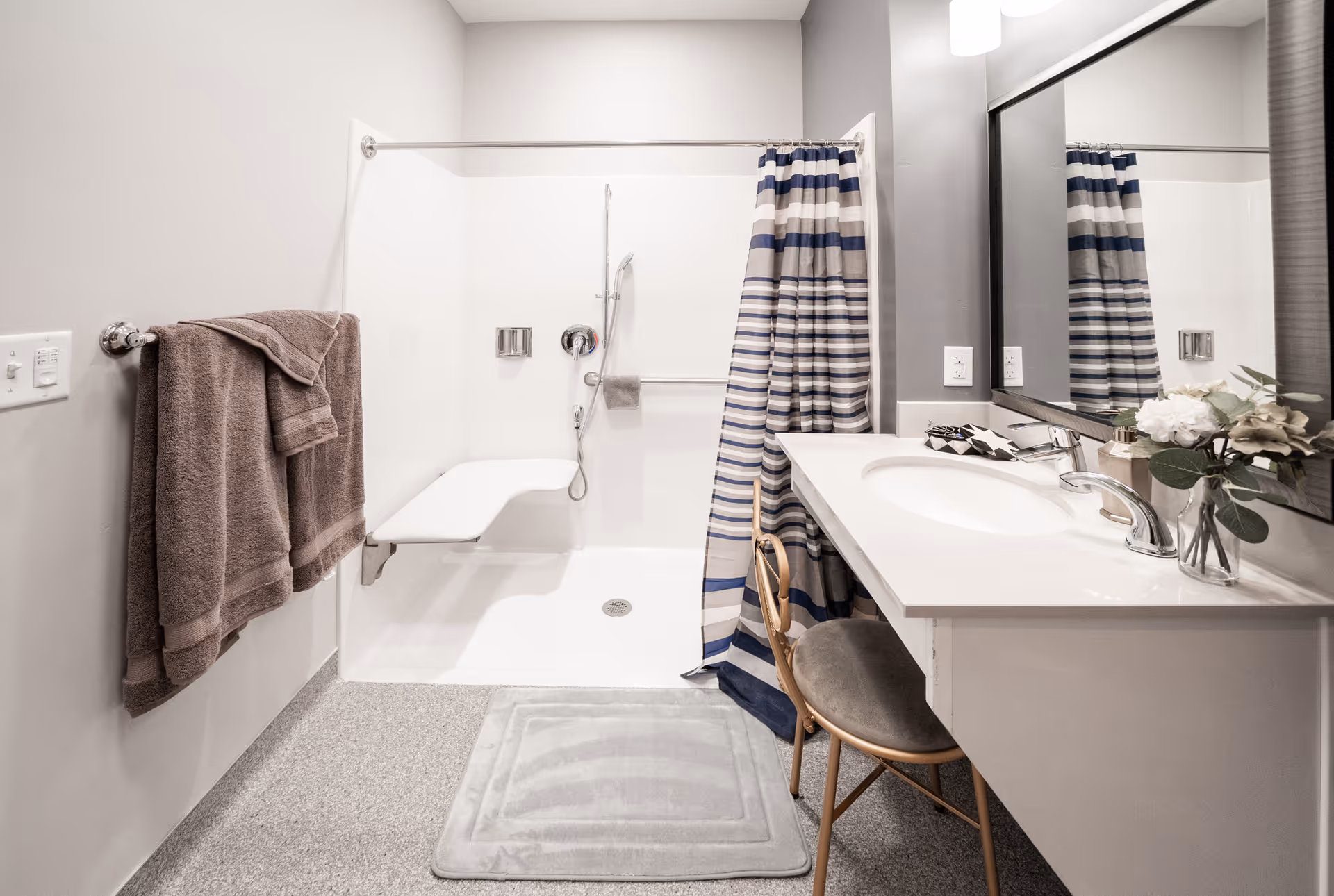 Accessible bathroom featuring a walk-in shower with a striped curtain, a vanity with sink and mirror, towels, and a small chair.