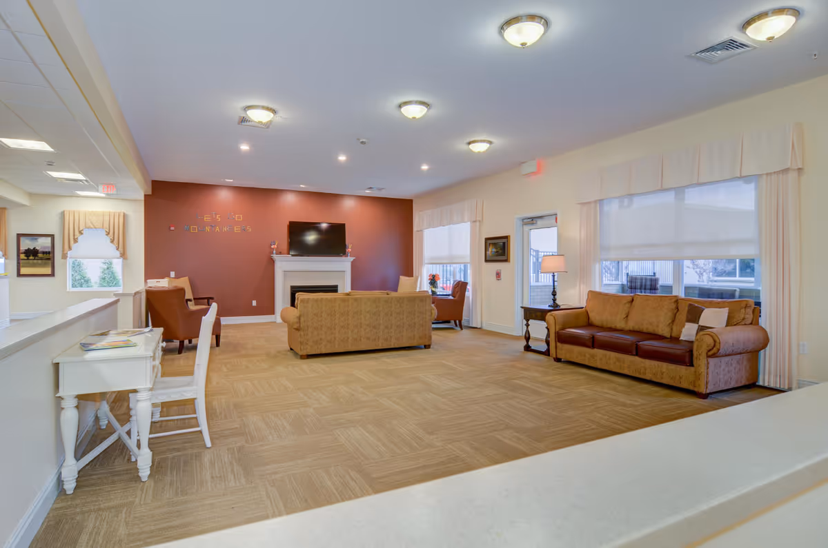 A spacious living room area in a senior living facility with beige carpet and cream-colored walls. The room features two sofas, two armchairs, a white fireplace with a flat-screen TV mounted above it, and large windows with white curtains. There is a small white desk with a chair in the foreground and a side table with a lamp next to one of the sofas. The far wall is painted a warm reddish-brown color with the words 'LET'S GO MOUNTAINEERS' displayed on it.