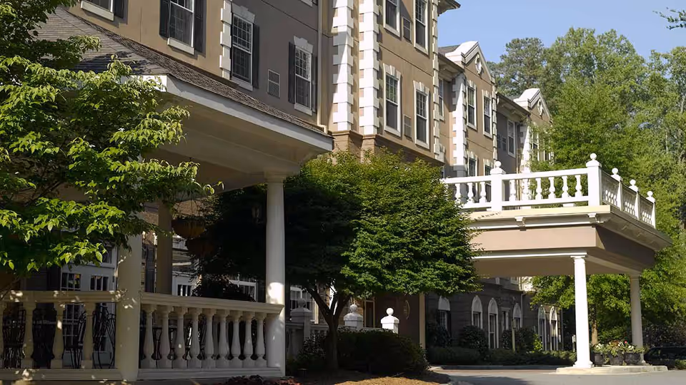 Exterior view of a multi-story senior living facility building with beige walls, white trim, and multiple windows. The entrance features a covered porch supported by white columns and a balcony above. There are green trees and shrubs around the building under a clear blue sky.