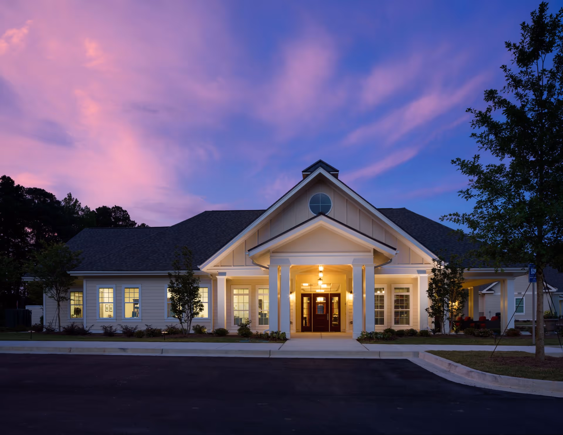 Front entrance of a single-story senior living community lit at dusk under a purple-pink sky.