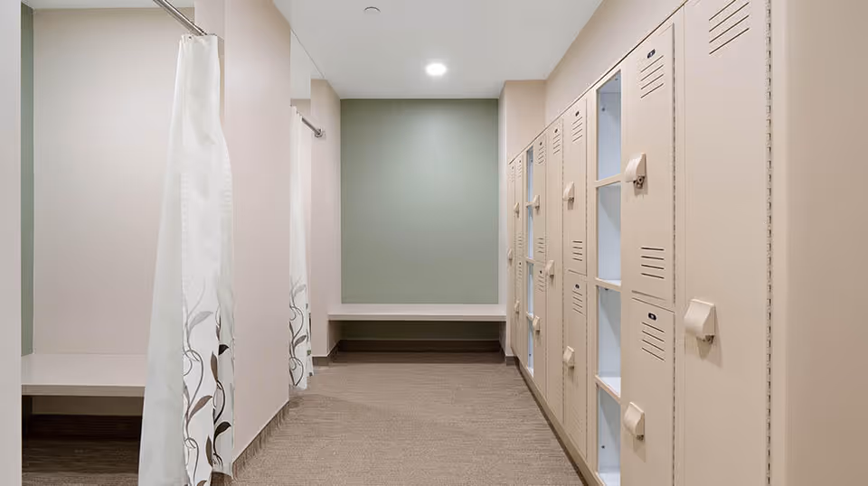 A clean locker/changing room with white lockers on the right, curtained changing stalls on the left, and a bench against the far wall.