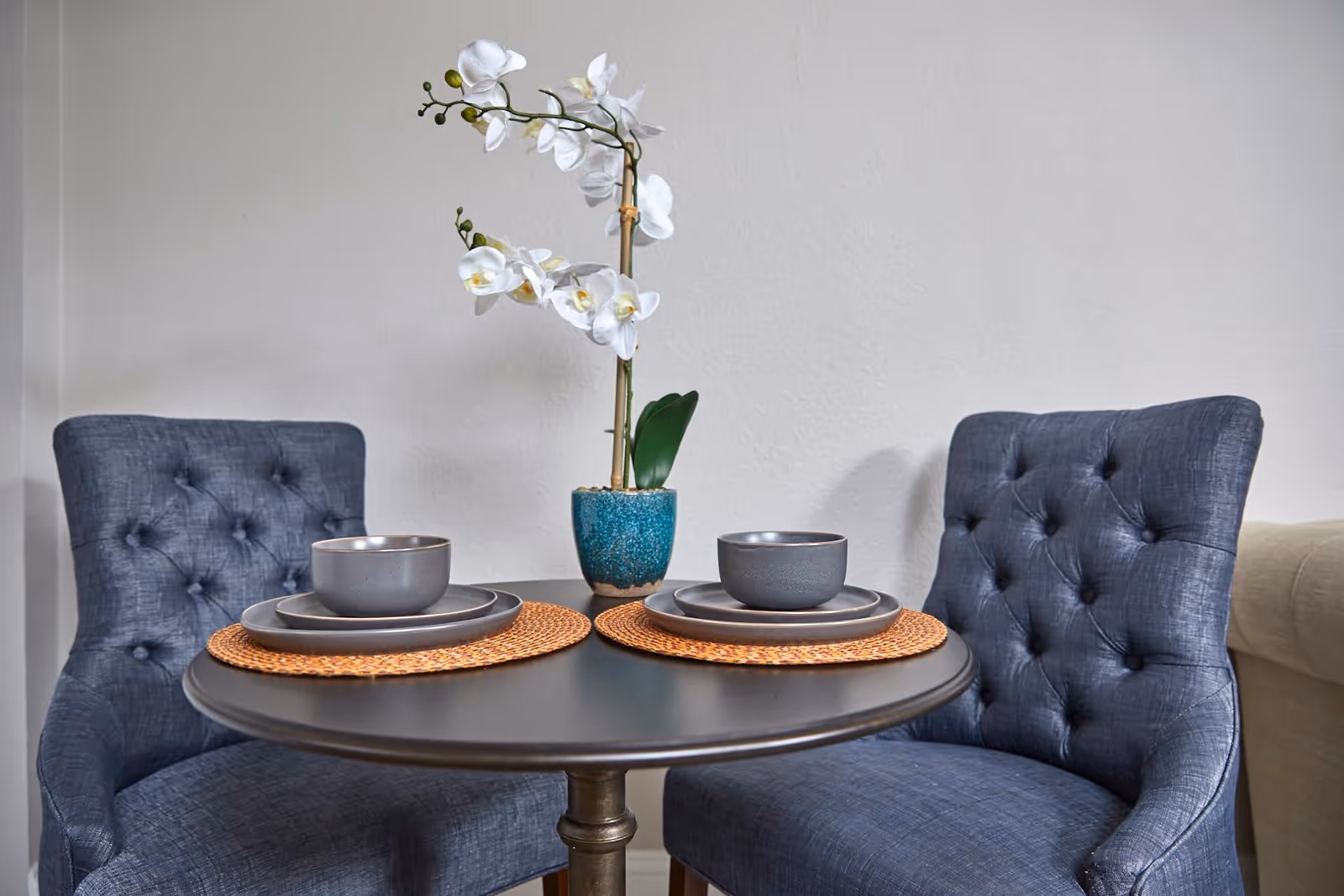 A small round dining table set for two with dark gray plates and bowls on woven placemats, flanked by two tufted dark blue upholstered chairs. A white orchid plant in a blue pot is placed in the center of the table against a plain light-colored wall.