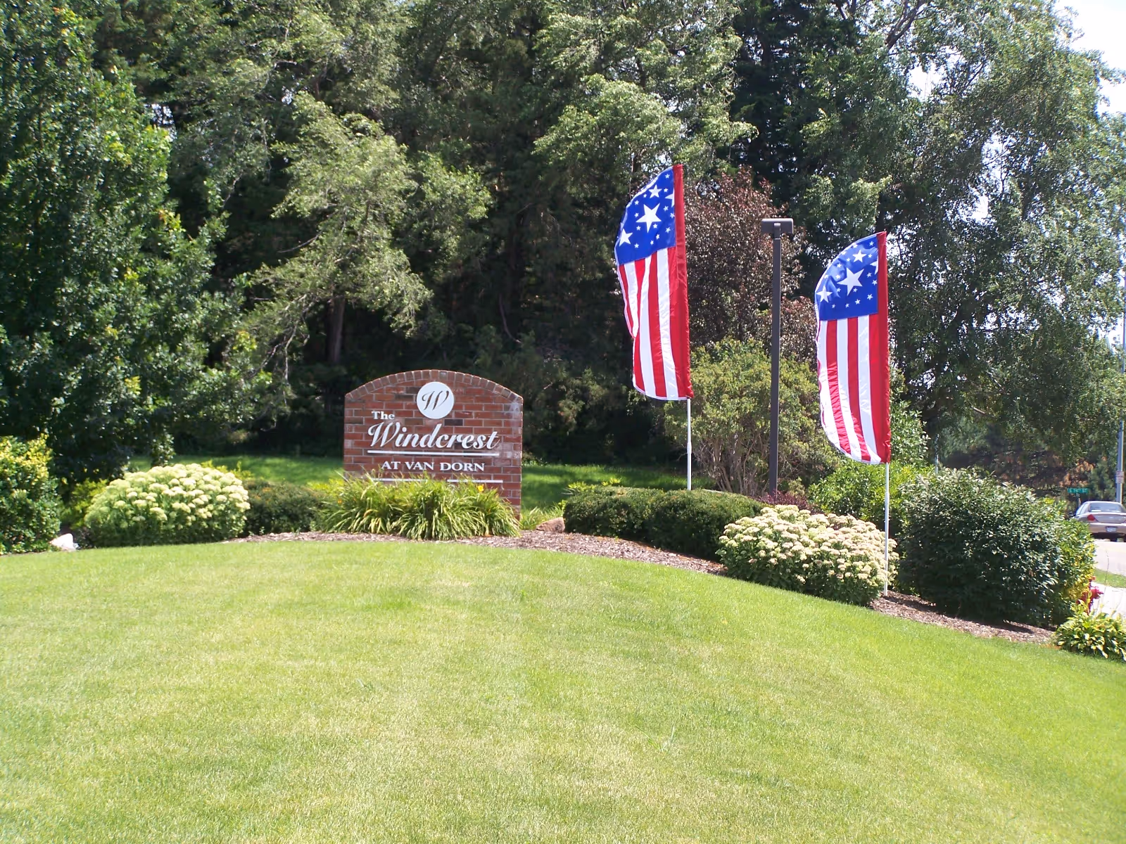 A landscaped outdoor area with a green lawn, bushes, and trees. There is a brick sign that reads 'The Windcrest at Van Dorn' and two American flags on poles beside the sign.