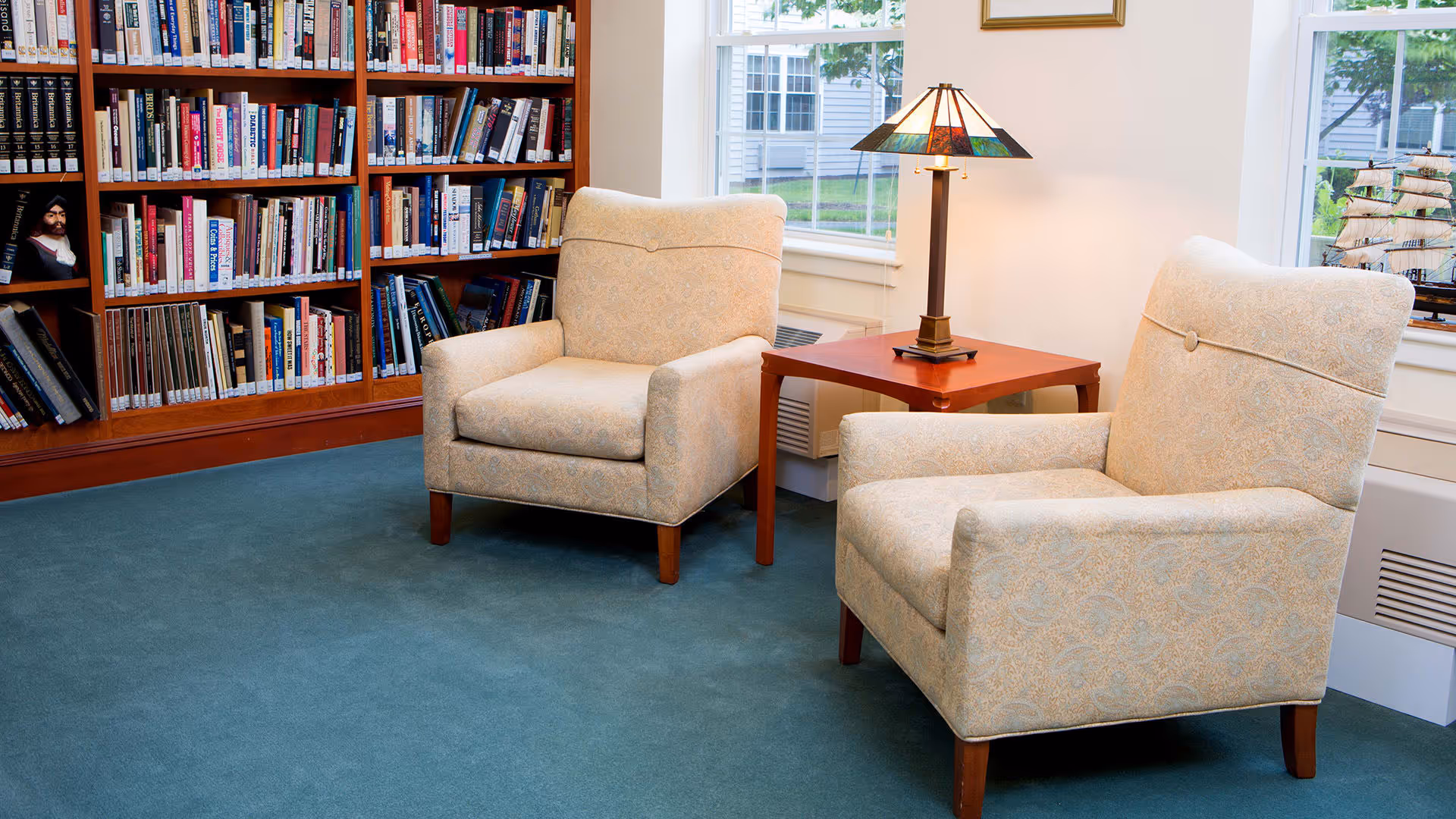 A cozy reading area with two beige patterned armchairs facing each other, separated by a wooden side table with a decorative lamp. Behind the chairs is a large wooden bookshelf filled with books. Two windows provide natural light and a view of greenery outside.