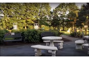 Outdoor garden seating area with stone picnic tables and benches surrounded by shrubs, trees, and decorative trellises.