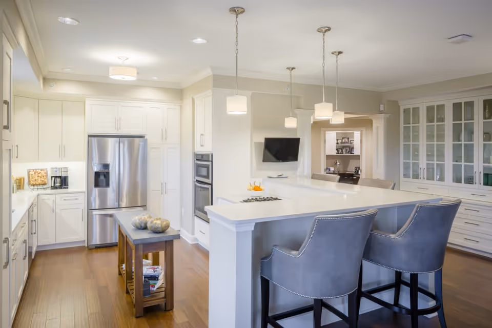A modern kitchen with white cabinetry, stainless steel refrigerator, built-in oven, and a large white island with a cooktop. Two gray upholstered bar stools are positioned at the island. The kitchen has wooden flooring, pendant lights hanging above the island, and a small wooden table with decorative items. In the background, there is a wall-mounted TV and a glass-front cabinet.