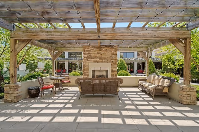 Outdoor seating area with a wooden pergola overhead casting shadows on the tiled floor. The area features a stone fireplace in the center, surrounded by wicker furniture including a sofa, loveseat, and chairs with cushions. Greenery and trees are visible in the background along with parts of the building.