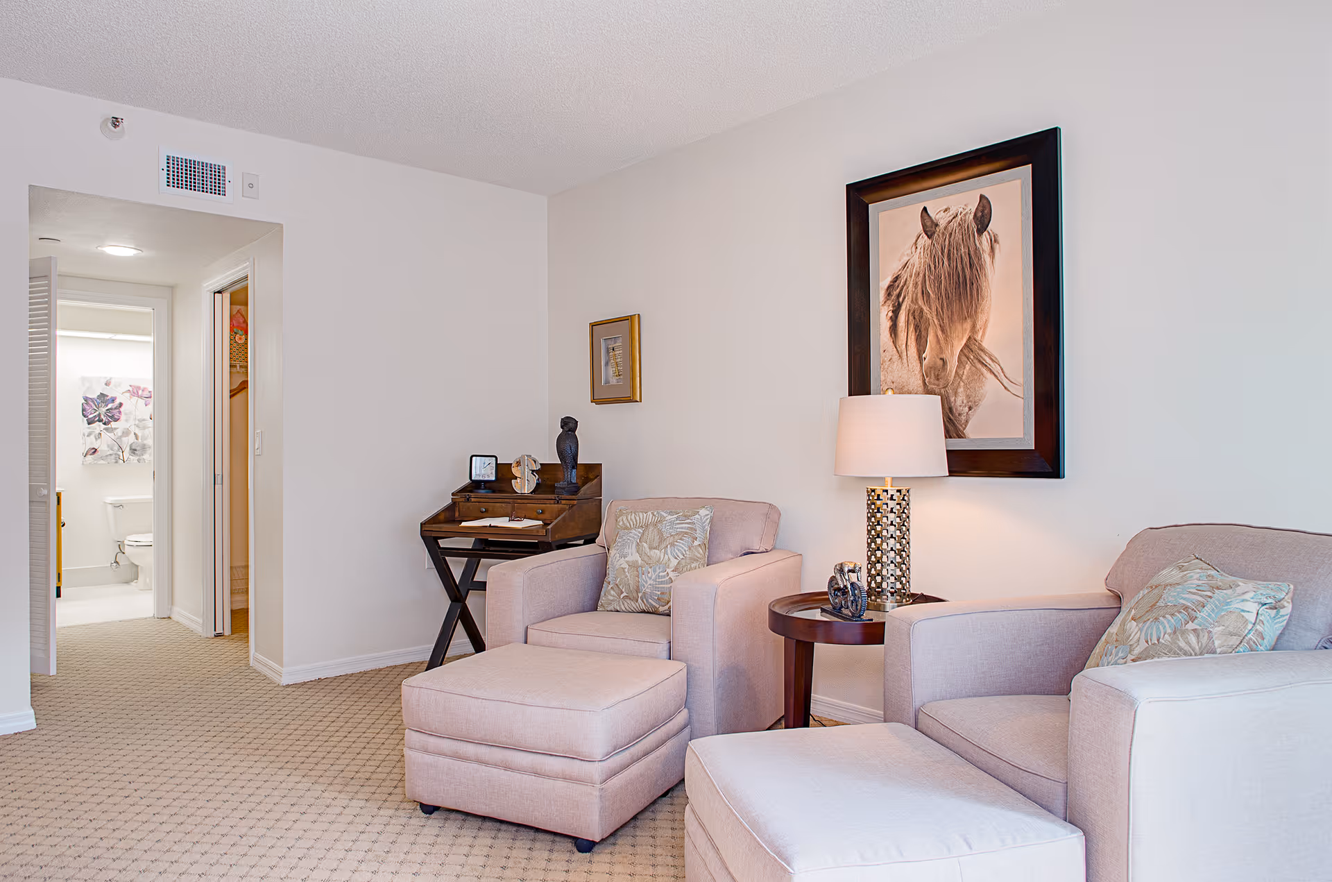 A cozy living room area with two beige armchairs and matching ottomans, a round wooden side table with a decorative lamp, and framed artwork of a horse on the wall. In the background, there is a small wooden desk with decorative items and an open doorway leading to a bathroom with a visible toilet and floral wall art.