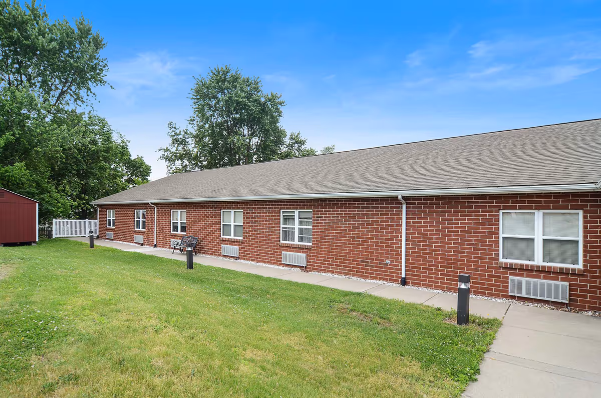 Exterior view of a single-story brick building with multiple windows, a gray shingled roof, and a concrete walkway alongside a grassy lawn. There are trees in the background and a small red shed on the left side.