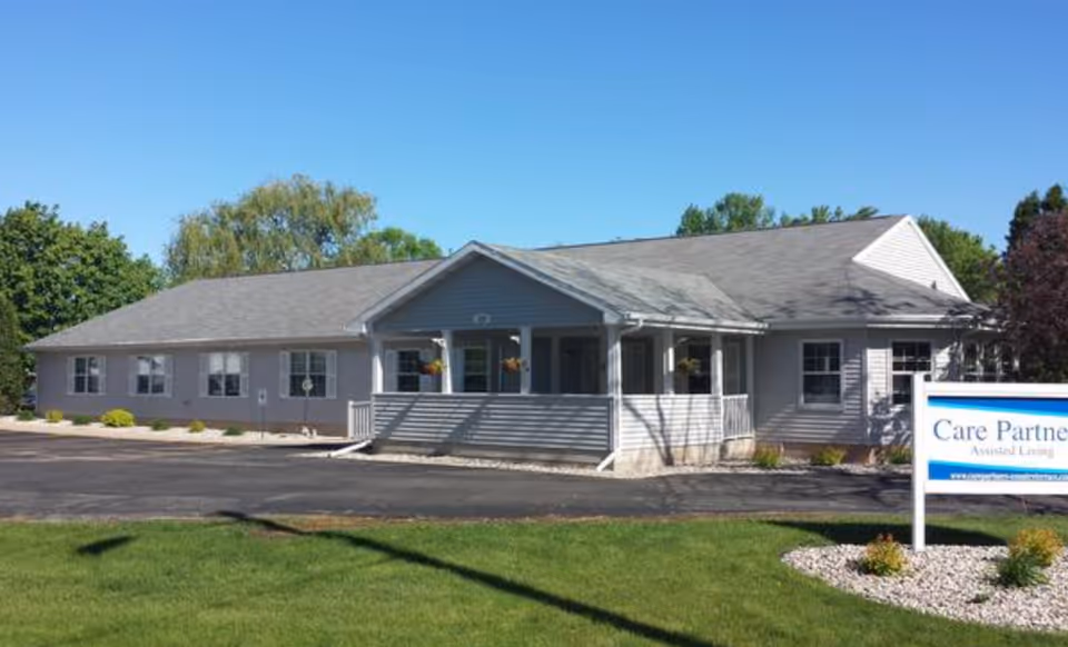 Exterior view of a single-story assisted living facility building with a gray roof and light-colored siding, a small porch with hanging flower pots, a paved driveway, green lawn, and a sign in front that reads Care Partners Assisted Living.