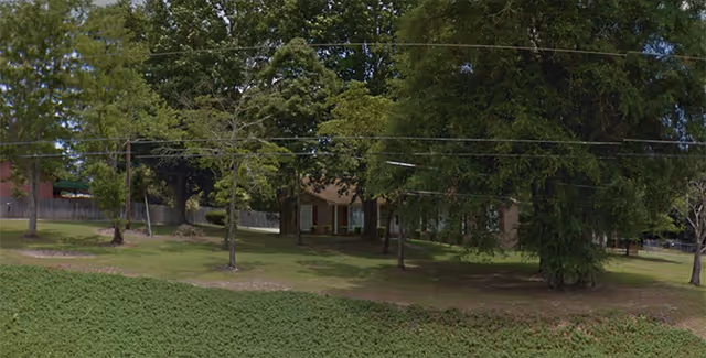 A grassy outdoor area with several trees and a building partially visible behind the trees. The scene appears to be a quiet, shaded space with some open lawn and a fence in the background.