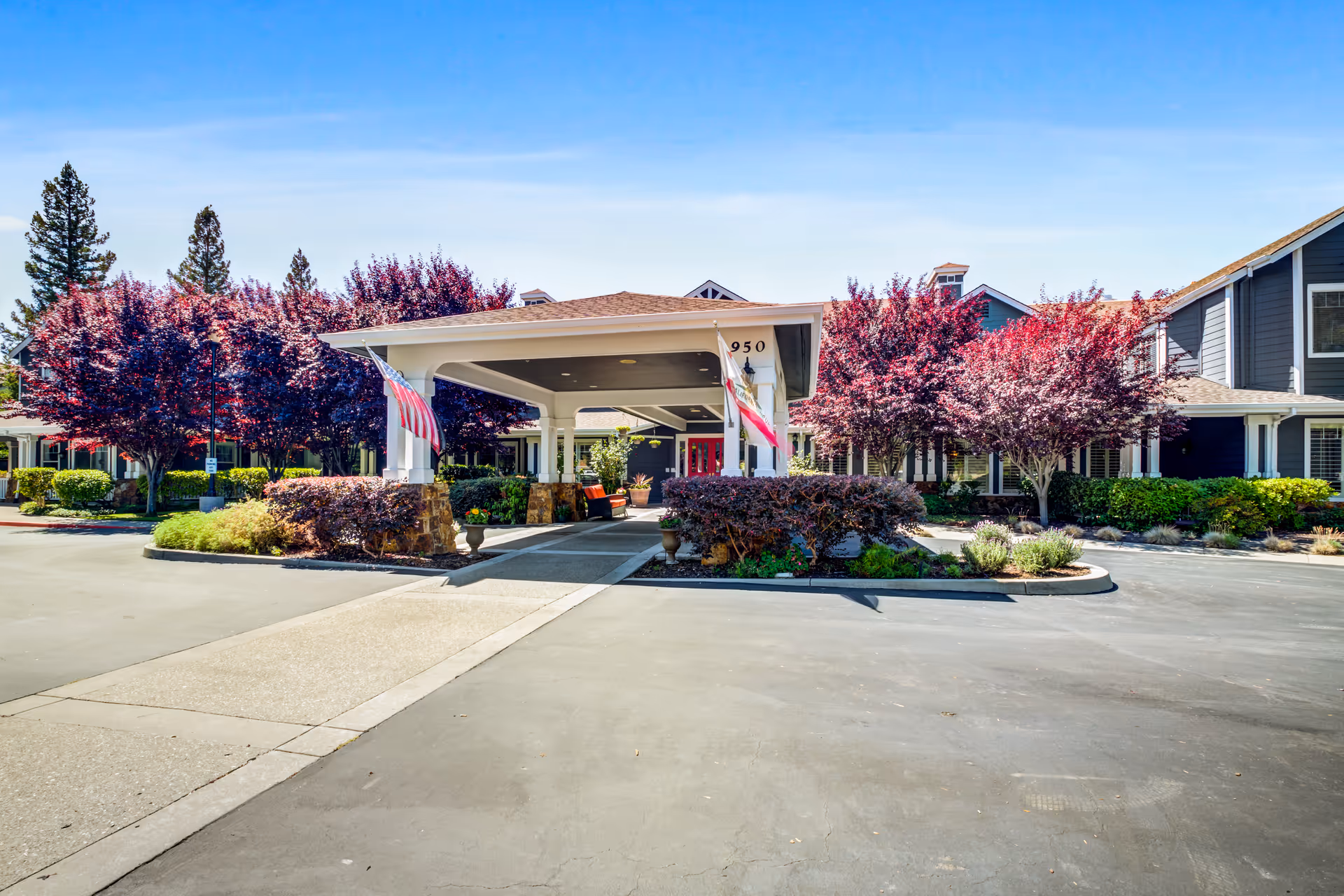 Porte-cochere entrance of a senior living building with American flags, landscaped planting beds and purple-leaved trees.