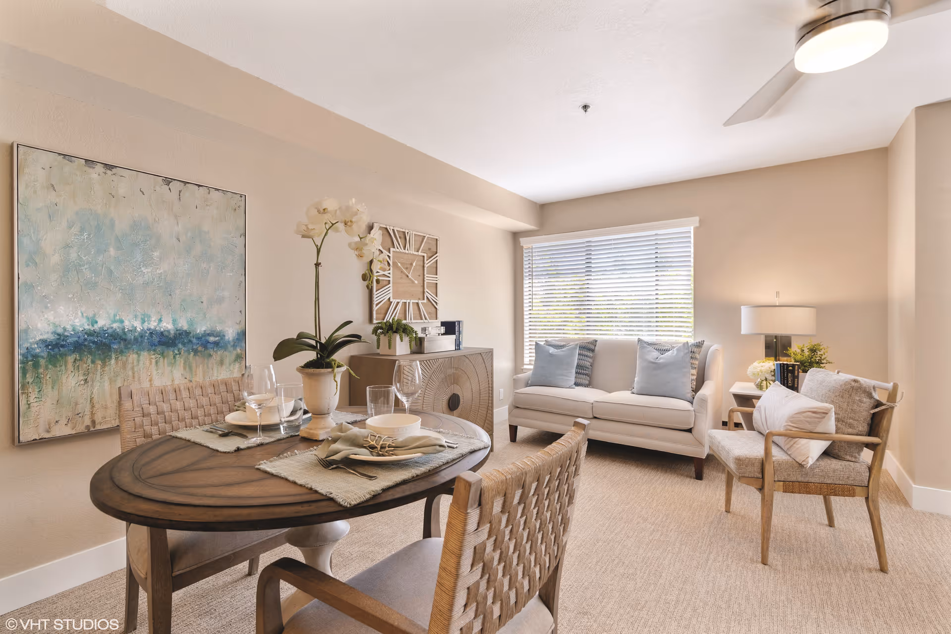 A cozy living and dining area with a round wooden dining table set for two, featuring woven chairs, a potted orchid centerpiece, and tableware. The room has a beige carpet, a white sofa with blue and patterned pillows, a wooden armchair with cushions, a side table with a lamp and books, a large window with blinds, a ceiling fan, a decorative wall clock, and an abstract painting on the wall.