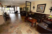 A spacious living room area with tiled flooring, featuring a dark brown leather sofa, matching armchairs, and a wooden coffee table with decorative items. The room has beige walls adorned with framed artwork and wall-mounted candle holders. Large windows and glass doors allow natural light to enter, and there are plants and additional seating near the entrance.