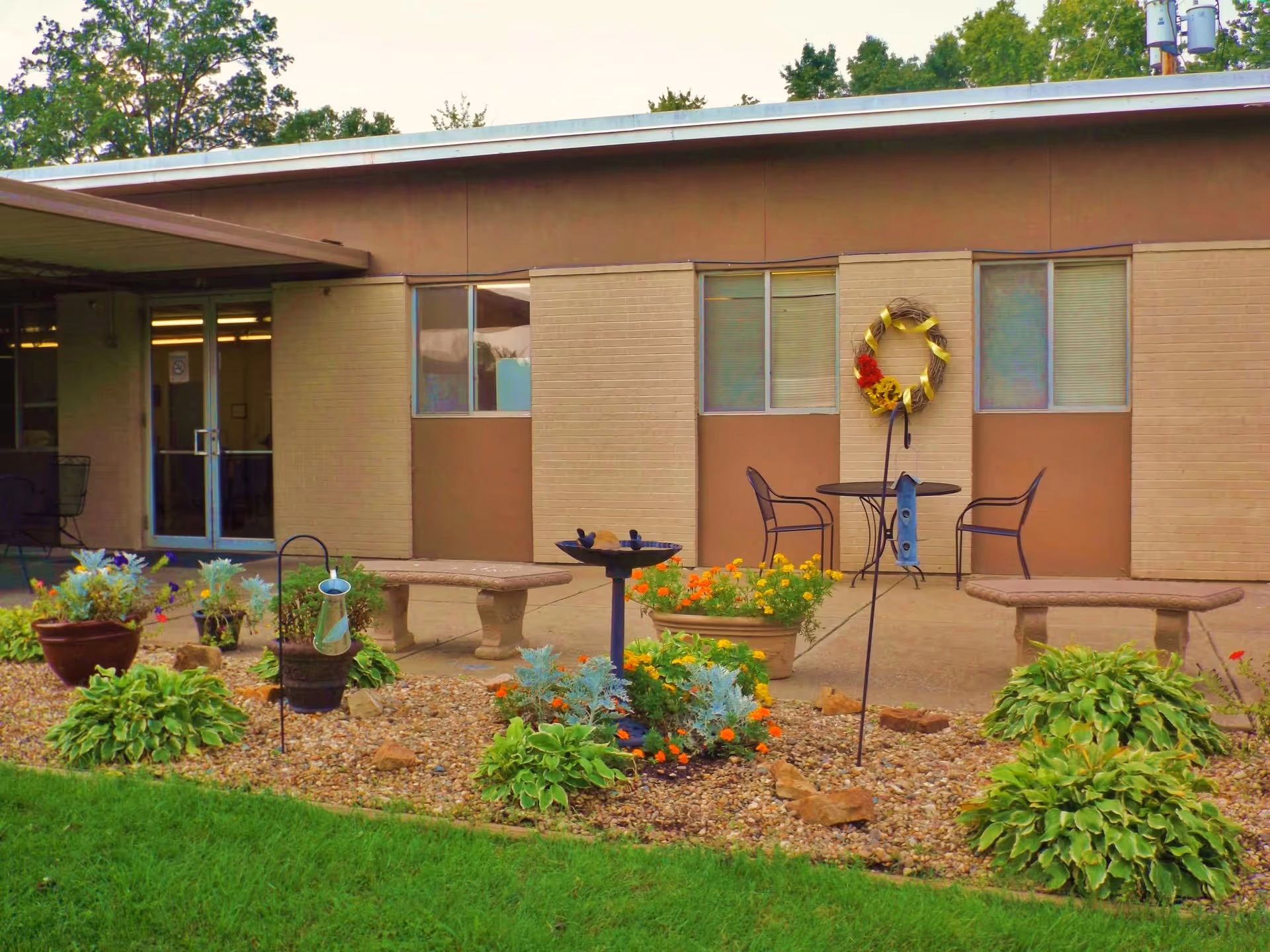 Outdoor patio area at a senior living facility with two stone benches, a round metal table with two chairs, a bird feeder, and a garden bed with various plants and flowers. The building exterior has beige brick walls and windows, with a decorative wreath hanging on the wall.