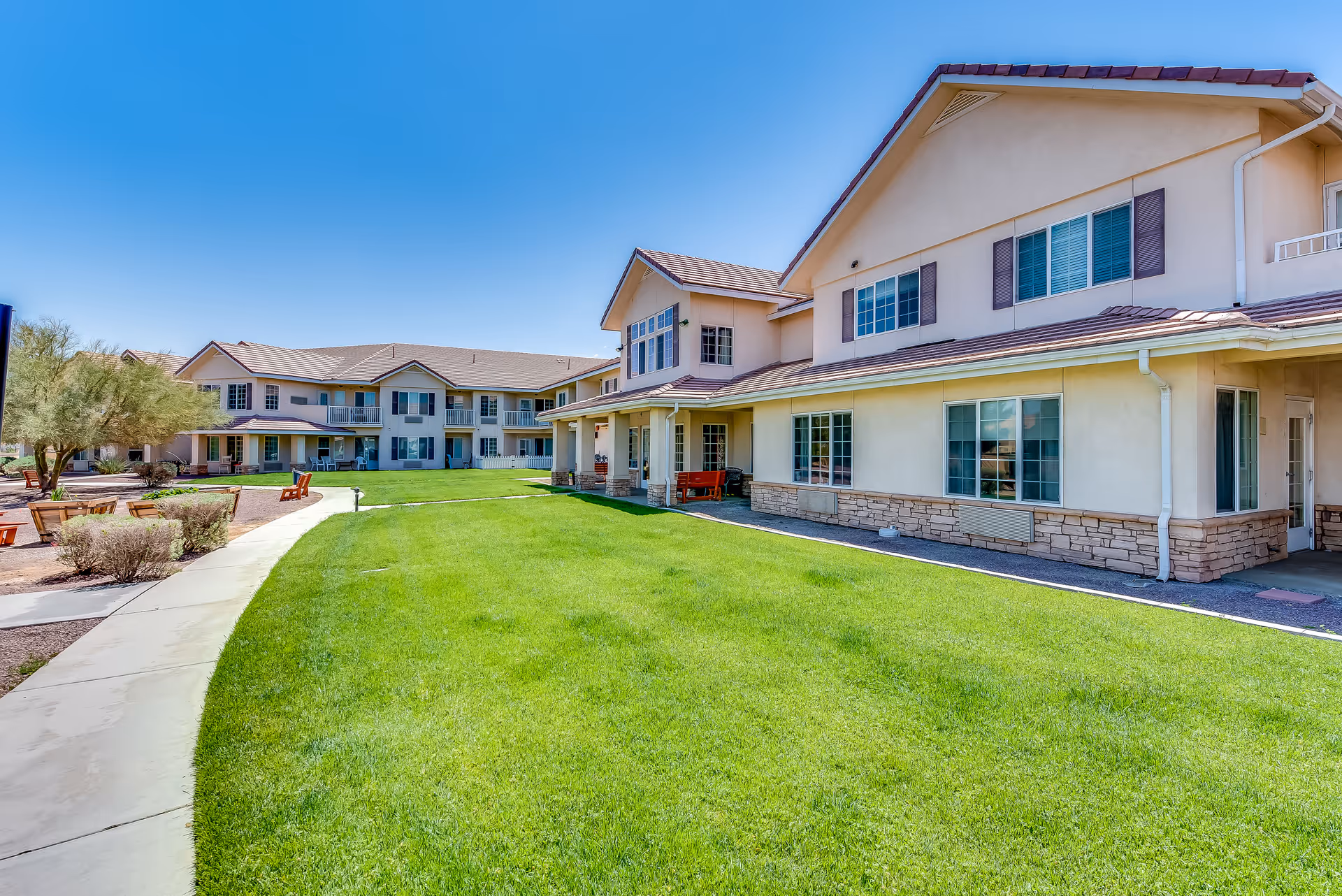 Two-story beige senior living building facing a green lawn with a curved walkway and outdoor seating under a clear blue sky.
