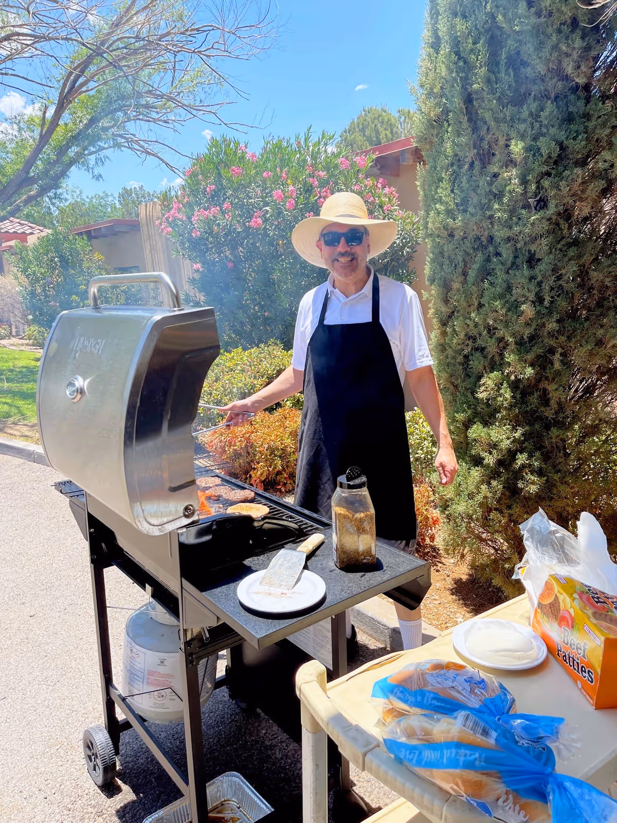 A man wearing a wide-brimmed hat, sunglasses, a white shirt, and a black apron is grilling food on a barbecue grill outdoors. There are bushes and trees in the background, and a table nearby holds hamburger buns, a package of beef patties, and plates.