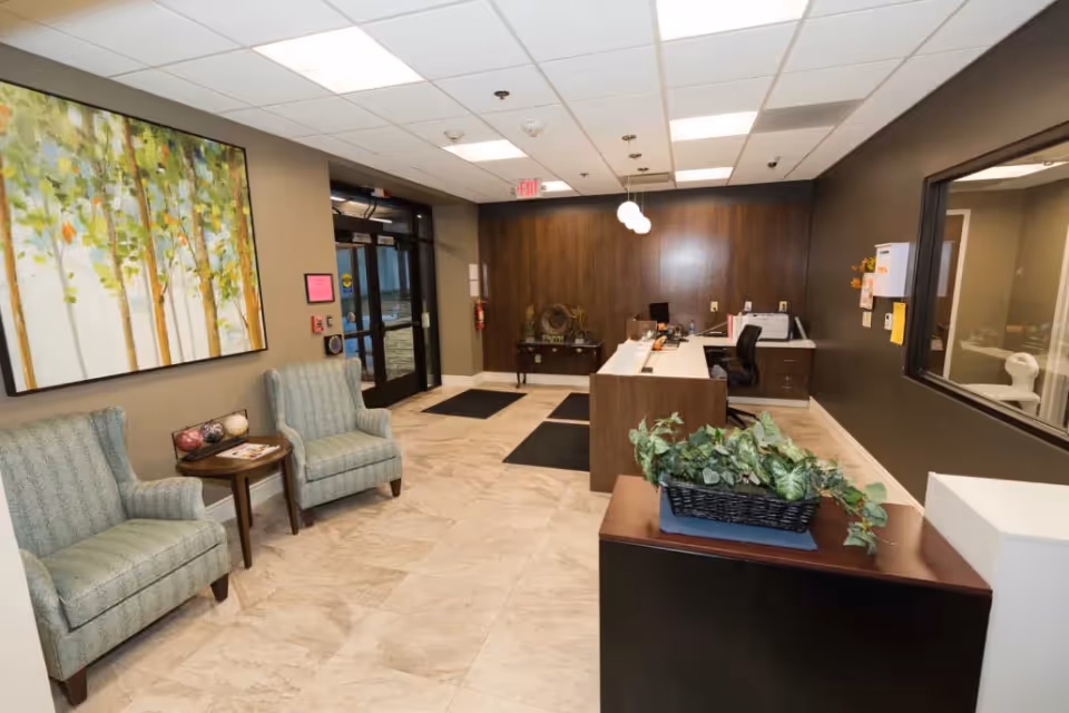Reception area of Keystone Ridge Post Acute Nursing and Rehabilitation featuring a front desk with office chairs, a small table with two upholstered armchairs, a large painting of trees on the wall, and a plant on a cabinet. The space has tiled flooring, overhead lighting, and a glass entrance door.