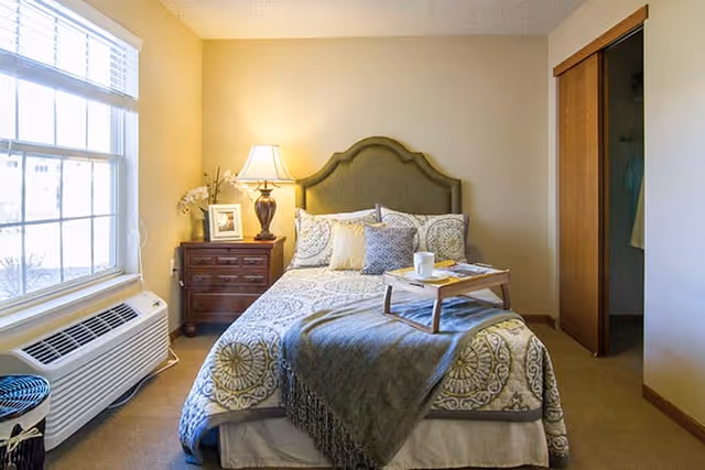 A cozy bedroom with a neatly made bed featuring patterned bedding and multiple pillows. A wooden nightstand with a lamp, a framed photo, and a small plant is beside the bed. There is a window with blinds on the left side, and an open closet with wooden sliding doors on the right. A small tray with a cup and some items is placed on the bed.