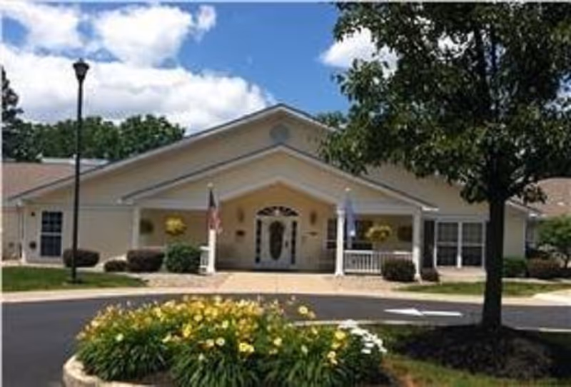 Front exterior view of a single-story building with a peaked roof, white columns, and a covered entrance. There are two flags near the entrance, a landscaped flower bed with yellow flowers in the foreground, a tree to the right, and a lamppost to the left under a partly cloudy blue sky.