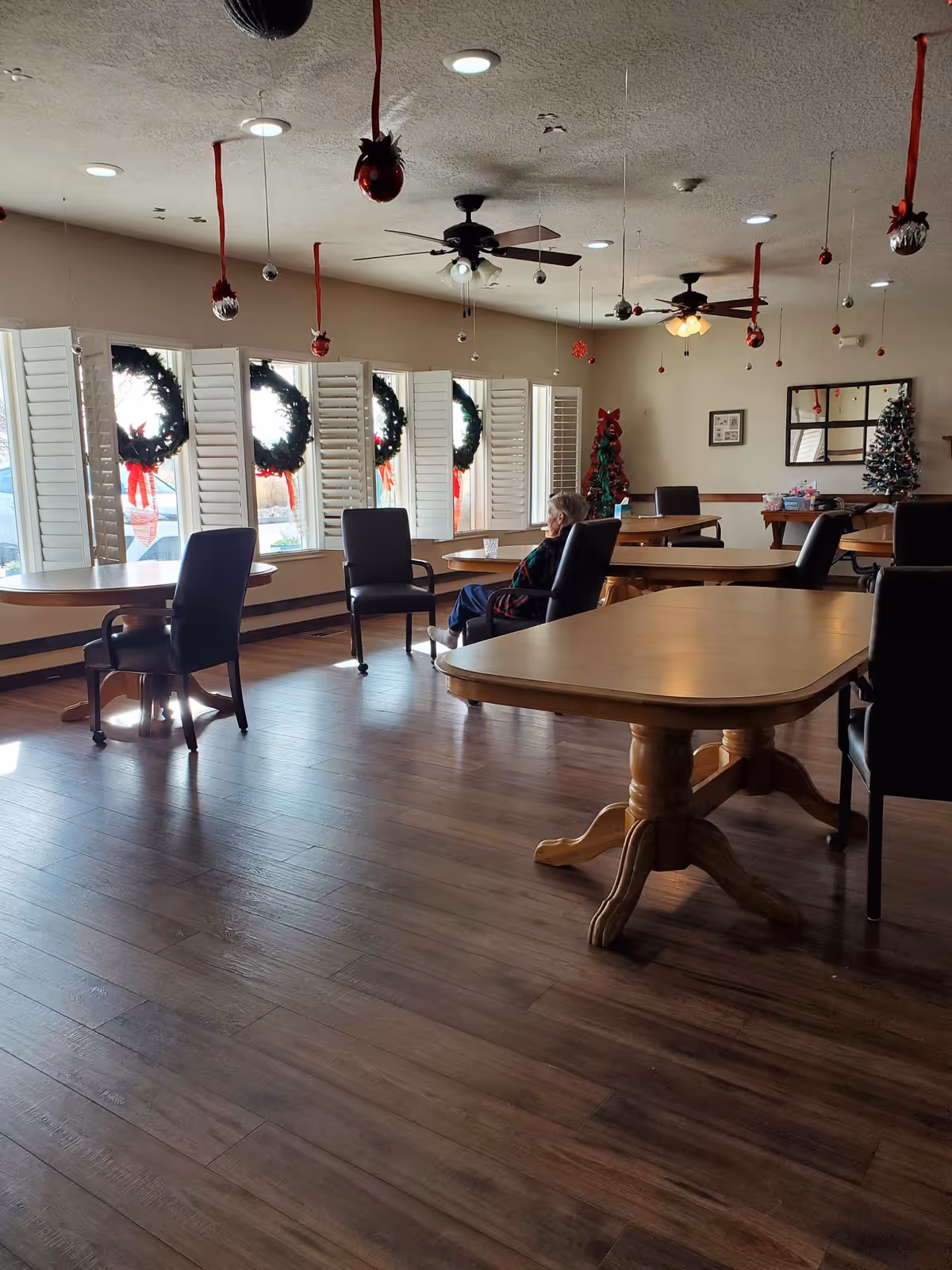 A spacious room with wooden floors and several wooden tables surrounded by black chairs. A single elderly person is seated near the windows, which are decorated with Christmas wreaths. The ceiling has fans and hanging Christmas ornaments, and there are two decorated Christmas trees in the background.