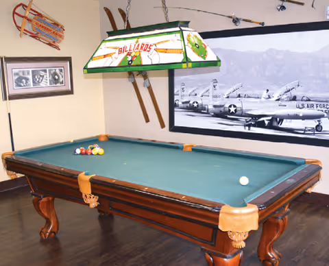 A billiards room featuring a green felt pool table with balls arranged for a game. Above the table hangs a stained glass light fixture with the word 'BILLIARDS' on it. The room has wooden flooring, vintage skis mounted on the wall, a framed black and white photograph of airplanes, and a sled mounted on the adjacent wall.