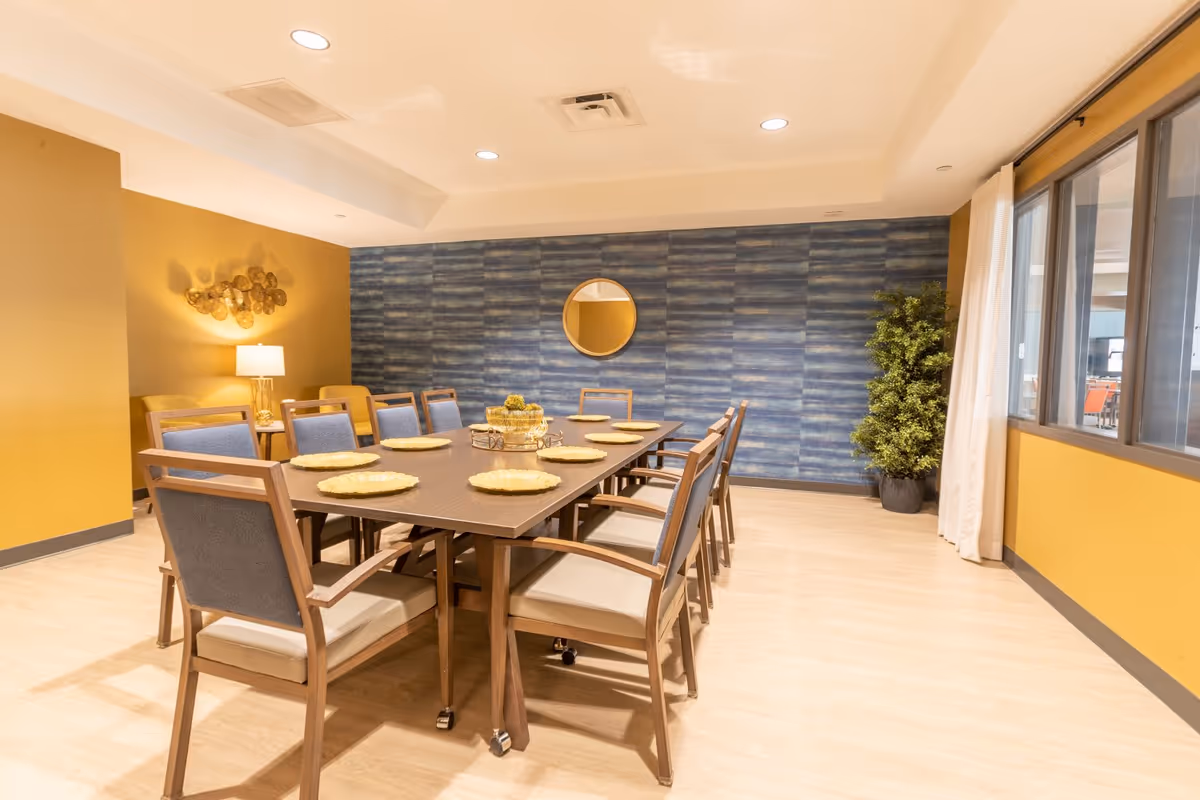 A well-lit dining room with a long rectangular table set with eight chairs. The table has decorative placemats and a centerpiece. The room features a blue textured accent wall with a round mirror, mustard yellow walls, a potted plant, a lamp on a side table, and large windows with white curtains.