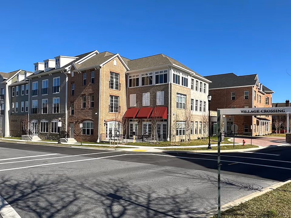 Front exterior of a multi-story brick and siding senior living building with a 'Village Crossing' entry archway at a street intersection.