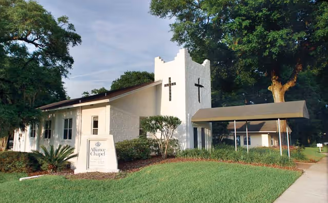 Exterior view of a white chapel building with two black crosses on the tower. The building is surrounded by green grass, bushes, and large trees. A covered walkway extends from the entrance to the sidewalk. A sign near the entrance reads 'Alliance Chapel'.