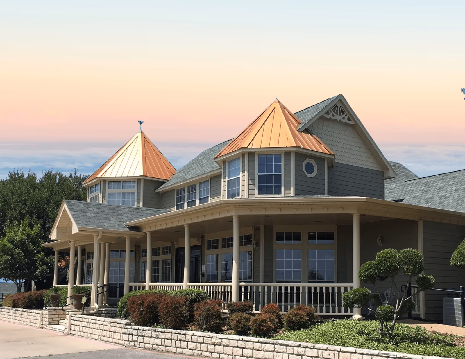 Front exterior of a two-story senior living building with a wraparound porch, copper-roofed turrets, and landscaped front yard.