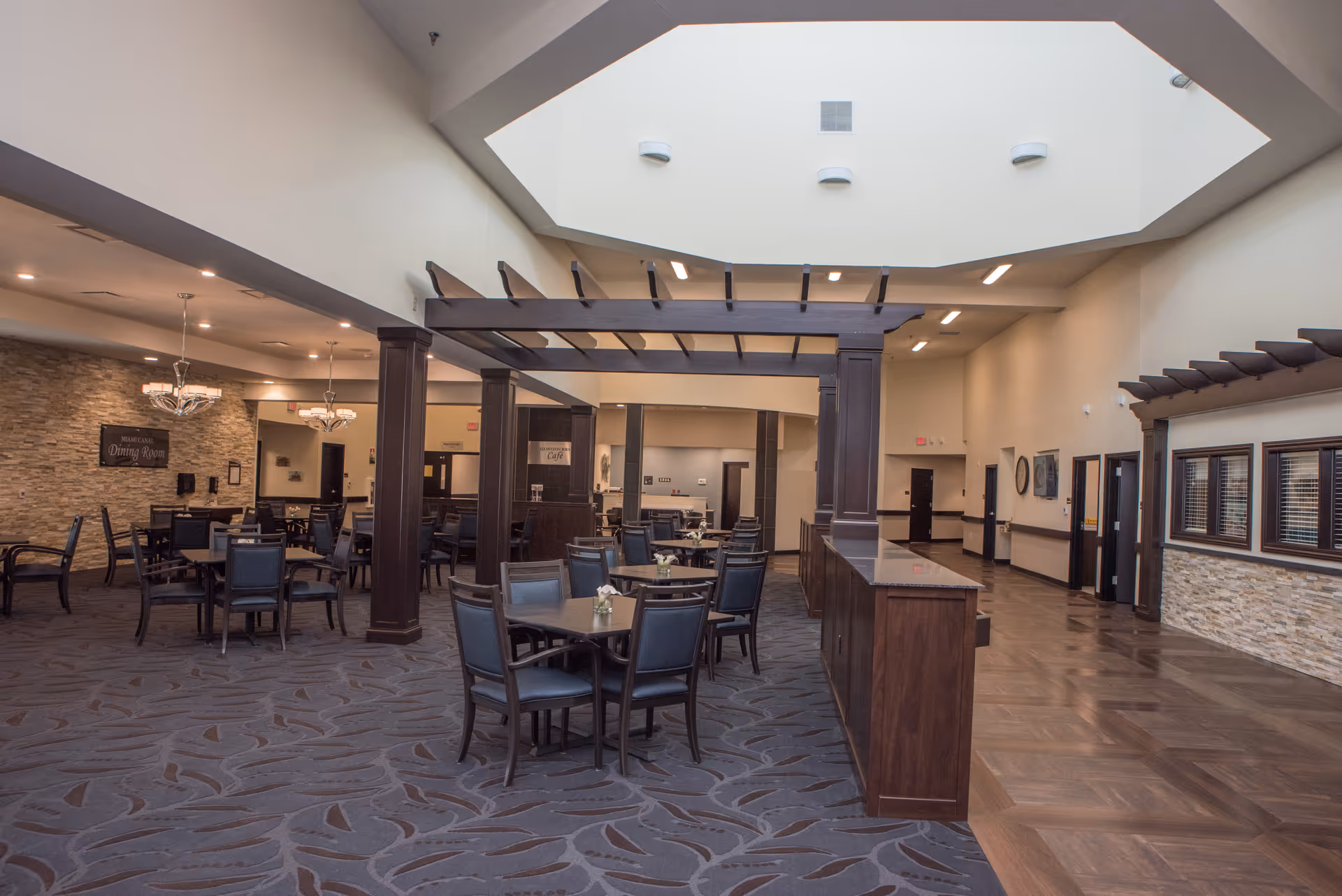 Spacious dining area with multiple tables and chairs arranged neatly. The room features a high ceiling with a large skylight, dark wooden beams and columns, and a combination of carpet and wood flooring. There are chandeliers hanging from the ceiling and a stone accent wall with a sign that reads 'Dining Room'.