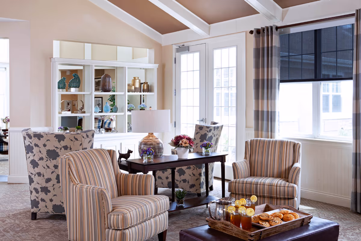 A cozy living room area with two striped armchairs and two floral-patterned armchairs arranged around a dark wooden coffee table. The table has a lamp, flower pots, and decorative items. Behind the seating area is a white shelving unit with various decorative objects. Large windows with striped curtains and a glass door allow natural light into the room. A tray with iced tea glasses and cookies is placed on a dark ottoman in the foreground.