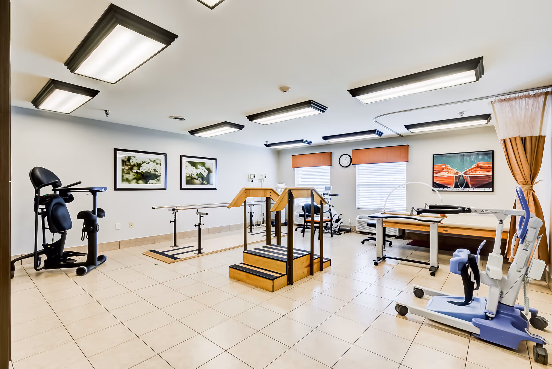 A spacious rehabilitation room with exercise equipment including parallel bars, a step platform with handrails, a stationary exercise bike, and a patient lift. The room has tiled floors, white walls with framed pictures, large windows with orange valances, and bright overhead lighting.