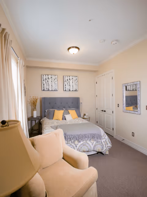 A cozy bedroom with a bed featuring a gray tufted headboard, patterned bedding, and yellow and gray pillows. There is a beige armchair in the foreground, a small side table with a vase of dried flowers, two framed black and white tree prints on the wall above the bed, a mirror on the right wall, and a closed double-door closet.