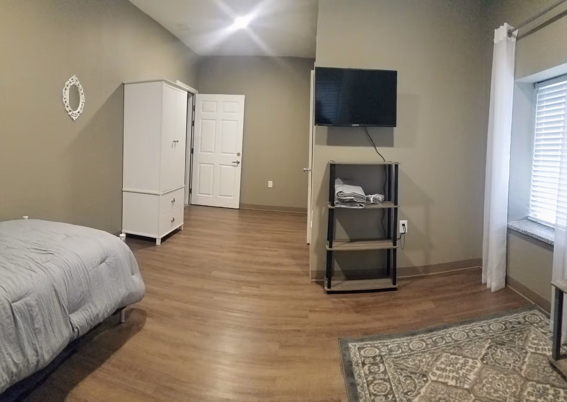 A simple furnished bedroom with a bed, white wardrobe, wall-mounted TV on a shelving unit, window and wood floors.
