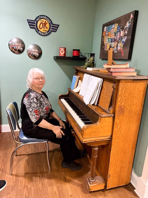 An elderly woman with white hair and glasses sits on a chair next to an upright wooden piano with sheet music and books on top. The room has light green walls decorated with vintage car signs and a colorful map of the United States made from license plates.