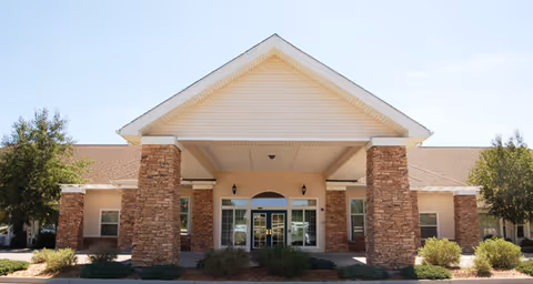 Front exterior view of a single-story building with a large covered entrance supported by stone pillars, surrounded by small bushes and trees under a clear blue sky.