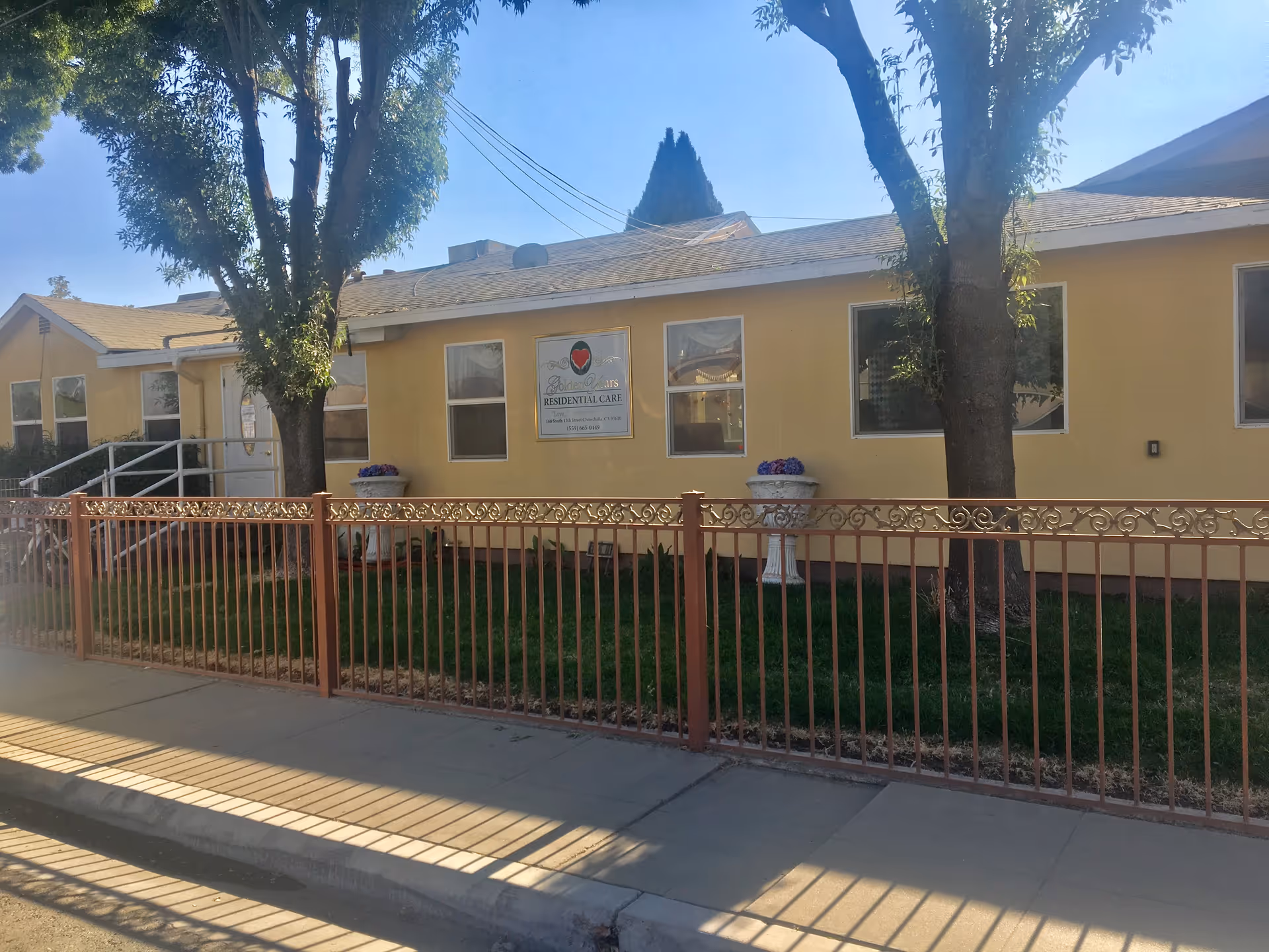 Exterior view of a single-story yellow residential care facility named Golden Years, with several windows, a metal fence in front, two large trees, and flower pots near the building.