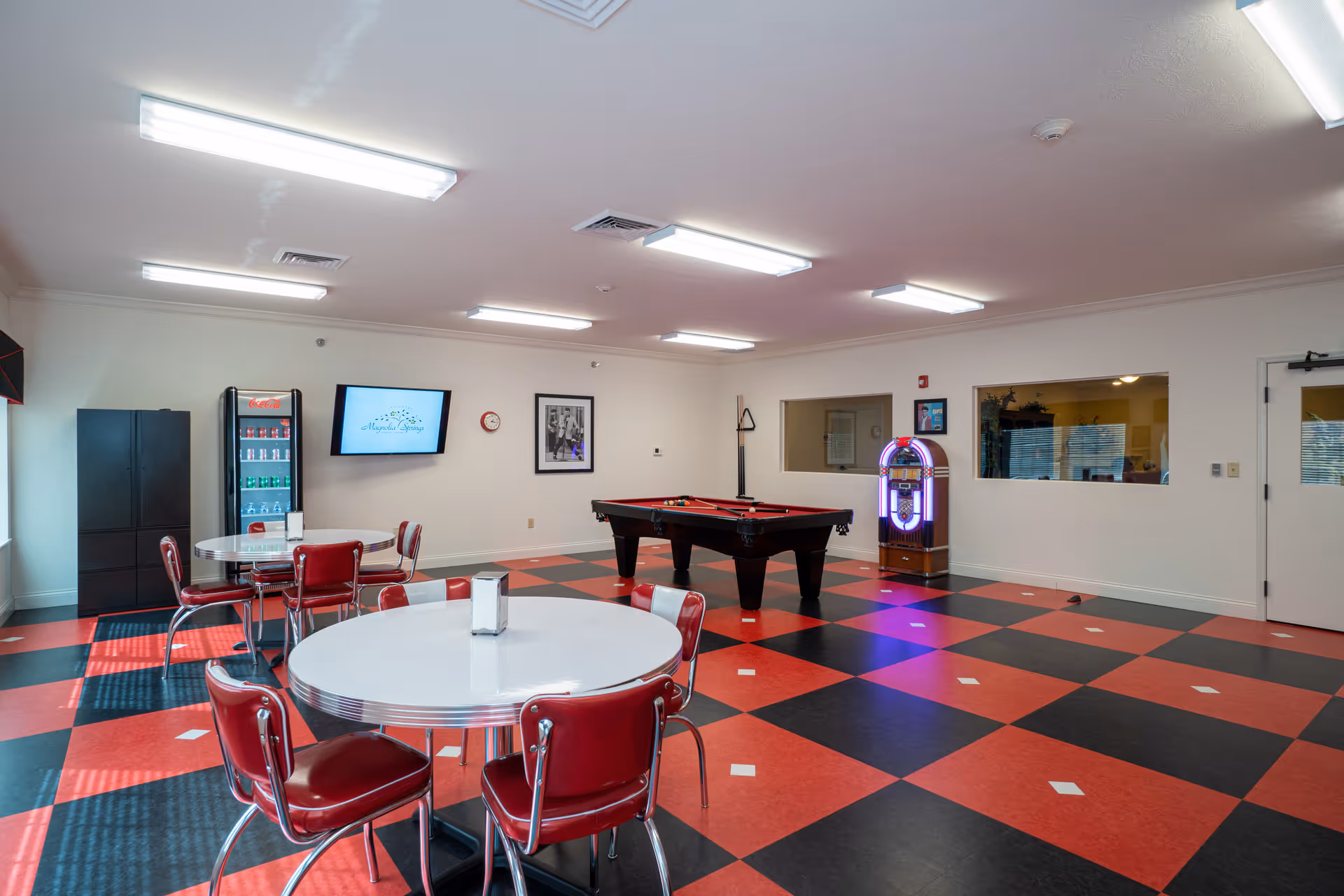 A recreational room with a red and black checkered floor, featuring two round white tables with red chairs, a pool table with a red felt top, a jukebox, a Coca-Cola vending machine, a black cabinet, a wall-mounted TV displaying the Magnolia Springs logo, and framed pictures on the walls.
