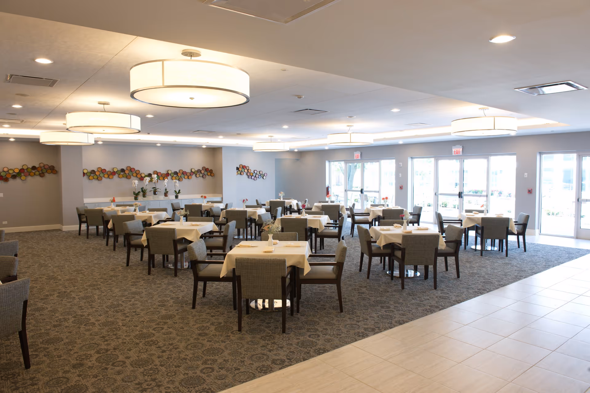 A spacious dining room with multiple tables covered in white tablecloths, each surrounded by four cushioned chairs. The room has large circular ceiling lights, carpeted flooring, and large windows letting in natural light. Decorative wall art and potted plants are visible along the walls.