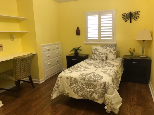 Small single bedroom with a twin bed dressed in patterned bedding, two dark nightstands, a desk and chair, a window with white shutters, and yellow walls.