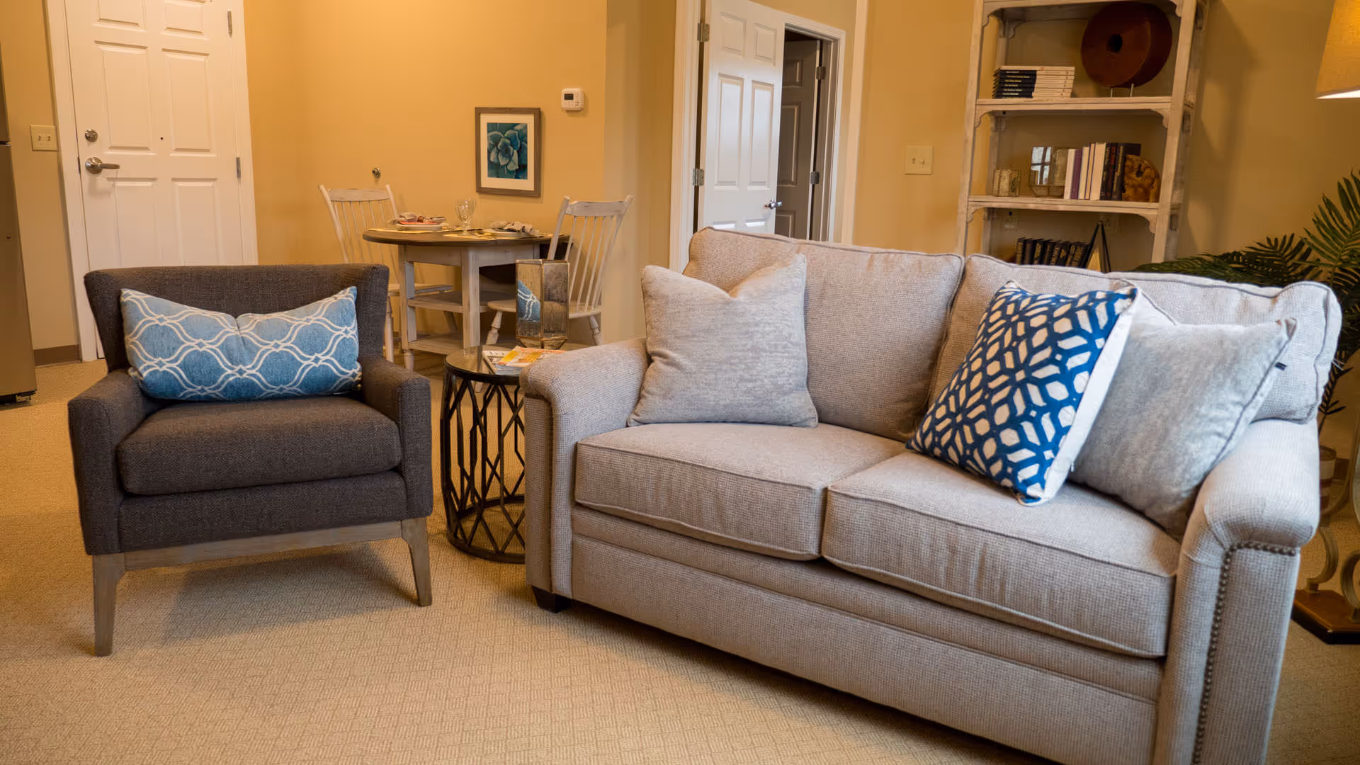 A cozy living room area with a beige sofa adorned with three decorative pillows in shades of blue and gray, a dark gray armchair with a blue patterned pillow, a round glass-top side table between them, and a small dining table with two chairs in the background near a white door. A bookshelf with books and decorative items is visible on the right side.