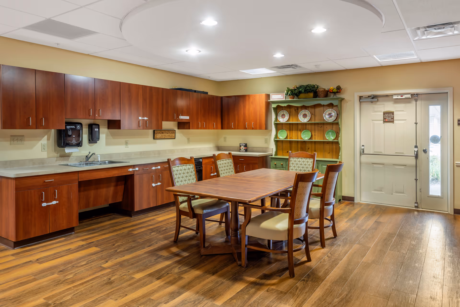 A kitchen area with wooden cabinets and a countertop along the wall, a sink, and a dining table with four chairs in the center. There is a green hutch with decorative plates and plants on top, and a door with a sign next to it. The floor is wood and the ceiling has recessed lighting.